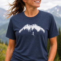 A woman wearing a navy blue heathered t-shirt with a white graphic of a mountain, pine trees, and a running person. The background shows a blurred outdoor mountain landscape.