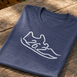 A folded heather navy blue t-shirt with a white outline graphic of a running shoe containing the text '26.2', displayed on a rustic wooden table.