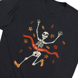 A close-up of a black t-shirt featuring a cartoon skeleton running with a red ribbon that reads 'for the season', surrounded by orange and yellow autumn leaves.