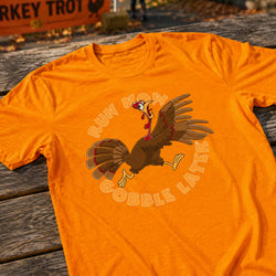 An orange short-sleeve t-shirt laid flat on a weathered wooden table, featuring a cartoon turkey graphic with the text 'RUN NOW GOBBLE LATER'.