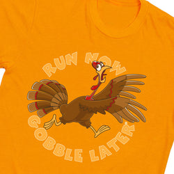 A close-up of an orange t-shirt featuring a cartoon graphic of a brown turkey with the text 'RUN NOW GOBBLE LATER' arched above and below in a light beige outline.