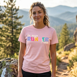 A woman wearing a light pink graphic t-shirt with the text 'in my RUNNER era' in colorful and white lettering, standing outdoors in a mountain forest setting.