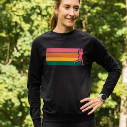 A woman wearing a black long-sleeve crewneck sweatshirt with a colorful retro striped graphic on the chest, standing outdoors in front of a green forest background.
