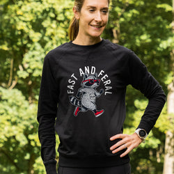 A woman wearing a black long-sleeve crewneck sweatshirt with a graphic of a raccoon wearing red sunglasses and red sneakers, with the text 'FAST AND FERAL' arched above it. She is standing outdoors in front of a blurred green forest background.