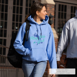 A woman walking outdoors wearing a light blue hoodie with a graphic that reads 'Play like a girl' in teal and dark blue. The photo is taken in a real urban environment with buildings and another person partially visible at the right edge.