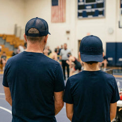 Two individuals wearing navy blue shirts and wrestling hats in a gymnasium setting.