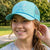 A smiling young woman outdoors wearing a light blue-green lacrosse trucker cap with 'LACROSSE' and crossed lacrosse sticks embroidered on the front. The background shows a blurred green park setting.