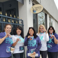 Five women in purple and white running shirts holding virtual race medals outside a cafe