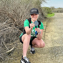 Female runner crouching on a trail holding a finisher medal and wearing a running hat and apparel
