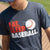 Boy at a field wearing a charcoal baseball t-shirt featuring red and white Eat Sleep Baseball slogan and flying ball graphic.