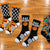 Six pairs of basketball-themed crew socks arranged on a polished wooden court floor. The designs include black and white web patterns, orange and black tiger stripes, black with 'EAT SLEEP' text and a basketball, a multicolor graffiti-style print, and a gray with a basketball graphic.