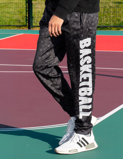 Youth wearing black basketball sweatpants featuring large vertical white Basketball text and subtle icon pattern.