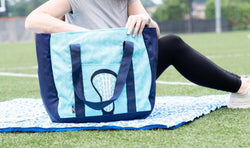 Light blue lacrosse tote bag with navy straps and a white lacrosse head graphic on a field.