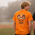 Two young men standing outdoors on a field, shown from the back. One wears a bright orange graphic T-shirt with a jack-o'-lantern bucket filled with candy, and the other wears a black graphic T-shirt with an orange skeleton character.