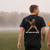 Two young men walking away on a misty grassy field, showcasing the back graphics of two different t-shirts. The man on the left wears a black t-shirt with a cartoon pumpkin graphic and the text 'HAT-TRICK OR TREAT'.