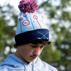Boy wearing light blue hockey knit hat with pom featuring an ice rink pattern design and embroidered crossed sticks on the cuff.