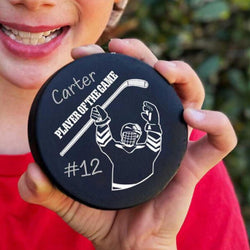 Personalized black hockey puck featuring Player of the Game text, custom name, and number held in hands.