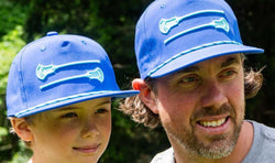 Father and son wearing matching royal blue lacrosse hats with a white crossed lacrosse sticks graphic.