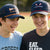 Two young men on a lacrosse field wearing caps and t-shirts with lacrosse-themed text.