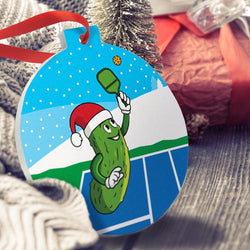 Pickleball Christmas ornament featuring a cartoon pickle in a Santa hat playing on a blue court.