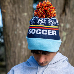 Boy wearing a navy and light blue knit soccer beanie featuring a multi-colored pom-pom and large Soccer text.