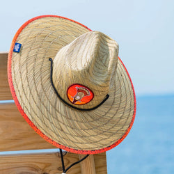 Straw hat with an orange embroidered lacrosse stick patch on the front, placed on the corner of a bench at a beach on a bright summer day with a blue sky in the background