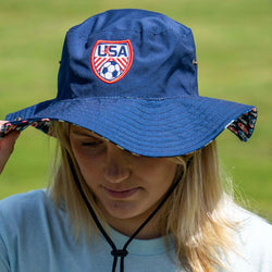 Person wearing a bucket blue hat with USA and soccer ball emblem on a grassy background