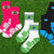 Four pairs of athletic crew socks arranged on a green grass background. The socks include pink and white, green and white, black and blue, and white and navy options, each with textured zones and football-themed graphics.