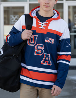 Boy wearing a navy blue USA hockey lace-up hoodie featuring red and white stripes and American flag graphic.