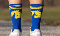 Blue crew socks with yellow unicorn graphic and stripes worn with white running shoes.
