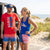 Two people in patriotic-themed apparel standing next to an open car door with a U.S. flag on a sandy path.