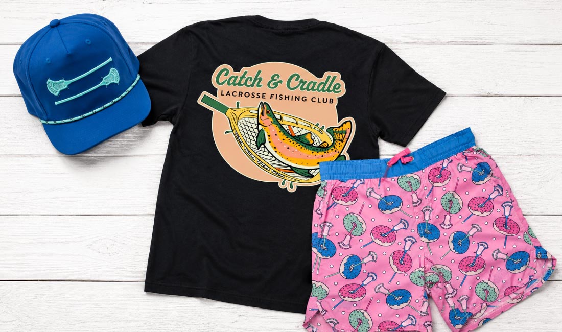 A flat-lay of an lacrosse themed outfit set featuring a black graphic t-shirt with a fishing club logo, pink shorts with a blue and green fish pattern, and a blue baseball cap with lacrosse stick graphics, all arranged on a white wood background