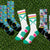 Six pairs of socks arranged on green grass. The socks feature various colorful patterns including cartoon dogs, crosscuts, and dog silhouettes on blue, white, pink, and black bases.