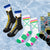 Four pairs of patterned and printed crew socks arranged in a row on a light blue ice texture background. The socks feature various designs including a black and yellow hockey theme, white with pink and green accents, and multicolor graphic prints.