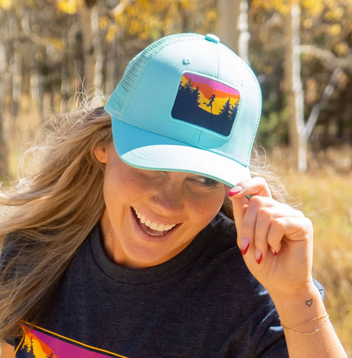 Light blue running trucker hat featuring a sunset trail runner graphic worn by a smiling woman outdoors
