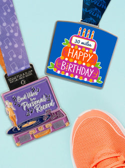 Two race medals and a running shoe on a light blue background. Top medal features a birthday cake with text 'HAPPY BIRTHDAY.' Bottom medal is shaped like a record player with text 'Good Vibes to a Personal Record.