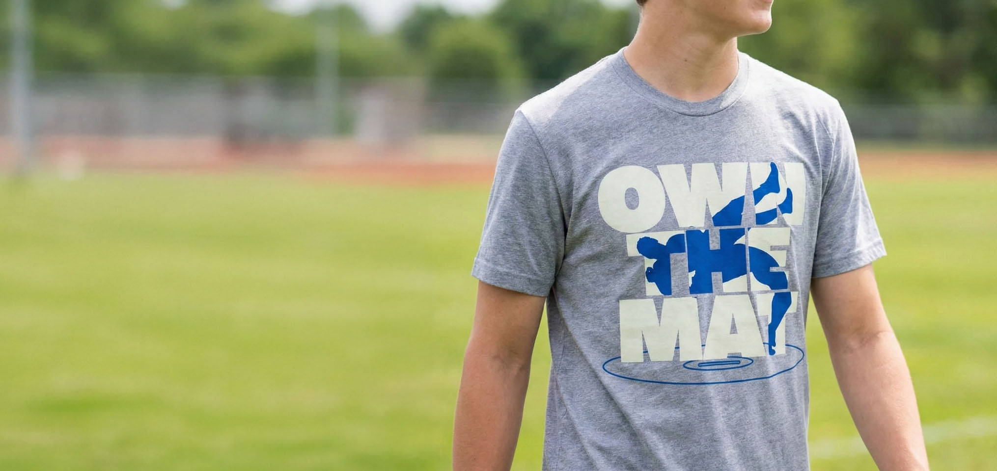 Youth wearing a gray wrestling t-shirt featuring Own The Mat slogan and blue wrestler silhouette.