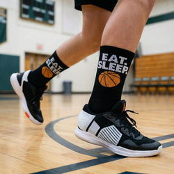 Person wearing black socks with 'EAT. SLEEP. BASKETBALL' and basketball graphics on a gym floor.