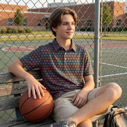 Young boy sitting on a bench with a basketball, wearing a patterned polo shirt and shorts, in an outdoor setting.