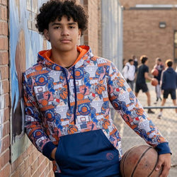 Person wearing a colorful hoodie with basketball-themed design, holding a basketball, standing against a brick wall.