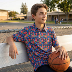 Boy in colorful basketball polo shirt holding a basketball, leaning against a fence outdoors.