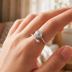 A silver ring with a baseball charm and a small bead accent, shown worn on a person's finger in a softly blurred indoor setting.