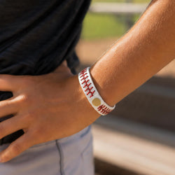 A close-up of a person's wrist wearing a white silicone bracelet designed to look like a baseball with red stitching and a gold patch.