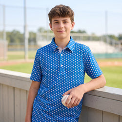 Young boy in a blue baseball patterned polo shirt holding a baseball on a sports field.
