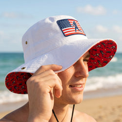Person wearing a white bucket hat with American flag and baseball player patch on a beach.