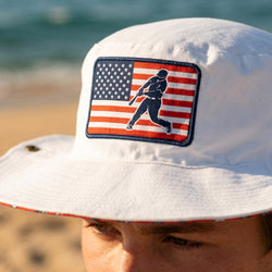 White bucket hat with American flag and baseball player patch on a beach.