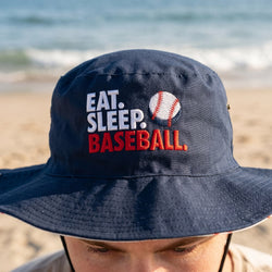 Navy blue bucket hat with 'Eat. Sleep. Baseball.' text and baseball graphic on a beach.