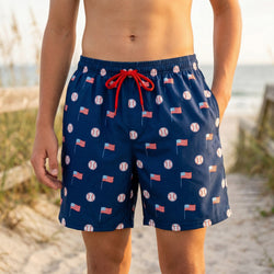 Navy blue swim trunks with baseball and usa flag pattern worn by a person on a beach.