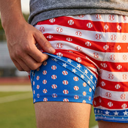 Person wearing red, white, and blue athletic shorts with baseball and star patterns on a blurred sports field background.