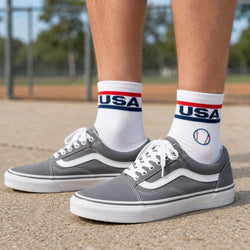 Gray Vans sneakers worn with white crew socks featuring a red, white, and blue USA design and a baseball graphic, shown on a person standing on a concrete surface outdoors.